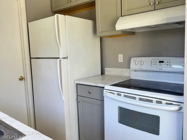Kitchen with gray cabinetry, white appliances, light stone countertops, and under cabinet range hood