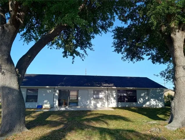Rear property exterior featuring a dark asphalt shingle roof, light-colored stucco walls, multiple windows, and a sliding glass door opening to a concrete patio