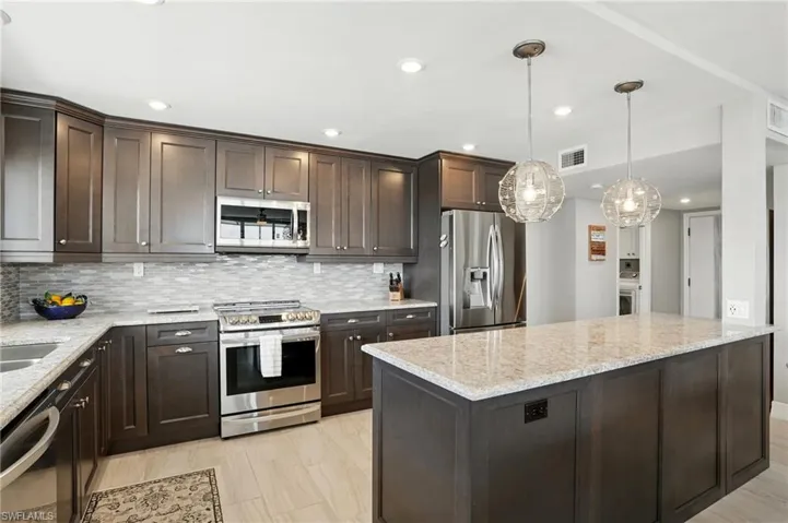 Kitchen with dark brown cabinets, appliances with stainless steel finishes, light stone countertops, hanging light fixtures, and recessed lighting - Virtually Edited Image