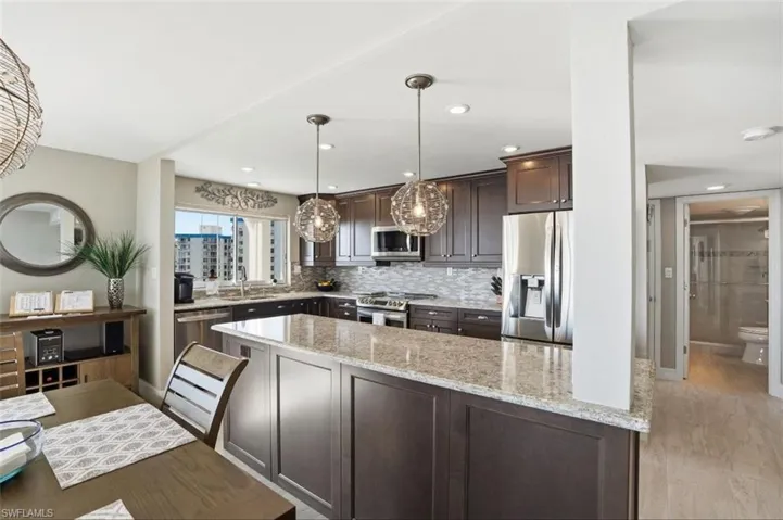 Kitchen featuring dark wood finish cabinets, stainless steel appliances, light stone countertops, decorative light fixtures, and tasteful backsplash - Virtually Edited Image