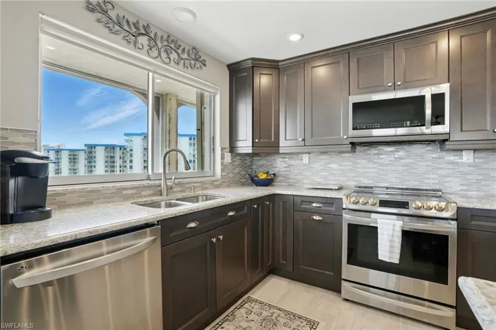 Kitchen with appliances with stainless steel finishes, dark brown cabinets, light stone countertops, backsplash, and a view of city - Virtually Edited Image