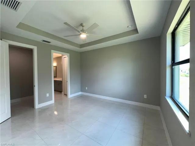 Unfurnished bedroom with light tile patterned floors, ensuite bath, ceiling fan, and a tray ceiling