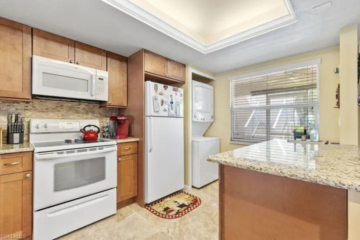 Kitchen featuring light stone countertops, white appliances, stacked washer / drying machine, tasteful backsplash, and light tile flooring