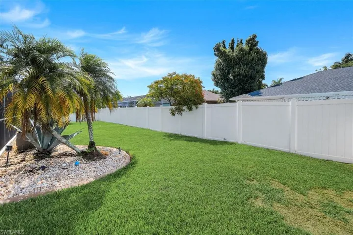 Expansive backyard featuring a lush green lawn, full white vinyl privacy fencing, and tropical landscaping with palm trees and decorative rock borders
