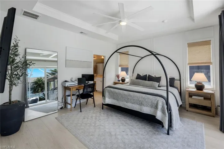 Bedroom featuring light wood-finish flooring, a tray ceiling with a ceiling fan, and large windows with natural fiber shades