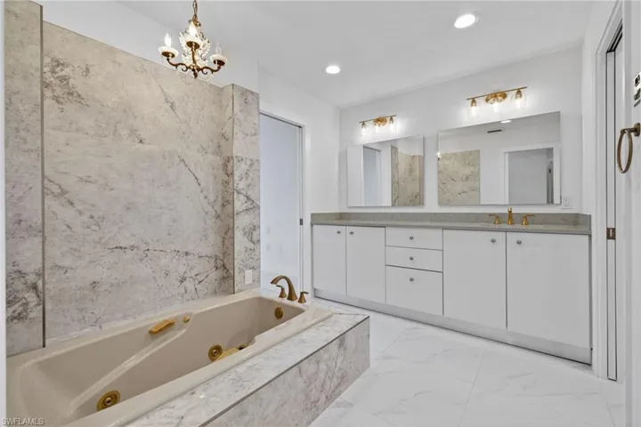 Full bath with a jetted tub, double vanity, light marble finish flooring, and suspended lighting