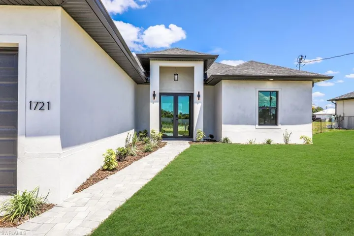 Modern stucco exterior featuring a covered entry with dual glass-paneled doors, a paver walkway, a dark shingle roof, and a mature lawn
