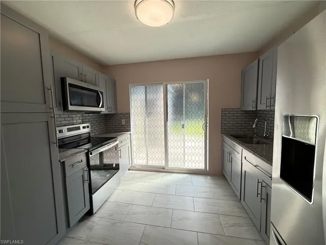 Kitchen featuring gray cabinetry, backsplash, stainless steel appliances, light marble finish floors, and dark stone countertops