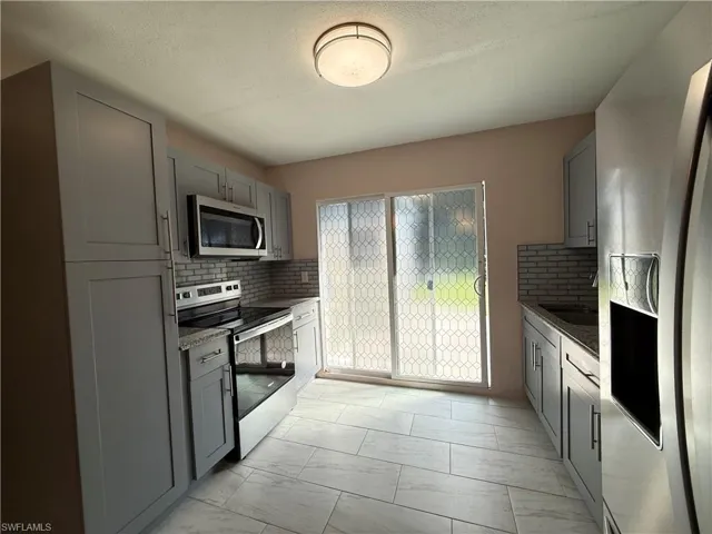 Kitchen with gray cabinetry, backsplash, stainless steel appliances, light marble finish floors, and light stone countertops