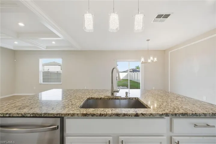 Kitchen featuring light stone counters, white cabinetry, coffered ceiling, stainless steel dishwasher, and suspended lighting