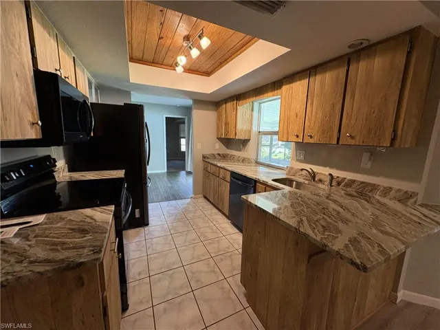 Kitchen with light tile patterned floors, sink, black appliances, a tray ceiling, and light stone countertops