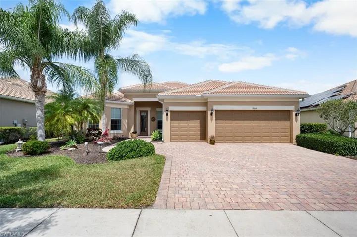 Beige stucco exterior with a tiled roof, featuring a paver driveway, two-car garage with panel doors, and tropical landscaping with mature palm trees