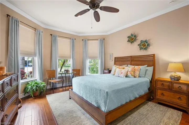 Bedroom featuring rich wood-finish flooring, a bay window with multiple panes, crown molding, and a ceiling fan with integrated lighting