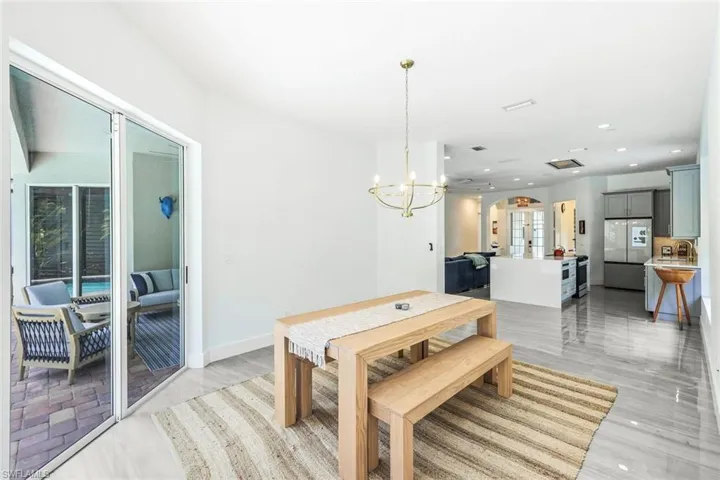 Open-concept living space featuring a dining area with a modern chandelier and light-toned flooring, opening to a covered patio with brick pavers
