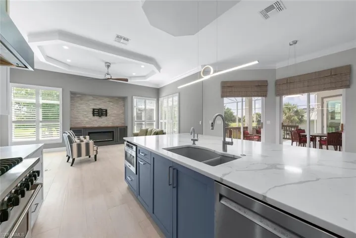 Open-concept kitchen and living area featuring a spacious island with a double basin sink, light-toned wood-finish flooring, a built-in fireplace with a stone surround, and multiple windows with plantation shutters