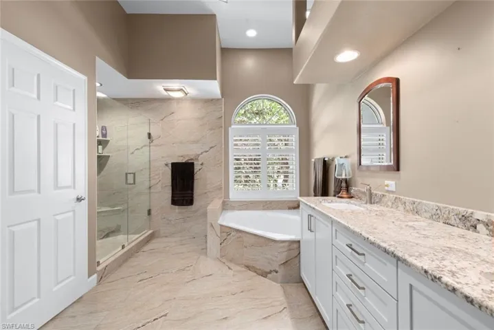 Spacious bathroom featuring a glass-enclosed shower, a corner built-in soaking tub, and an arched window with plantation shutters