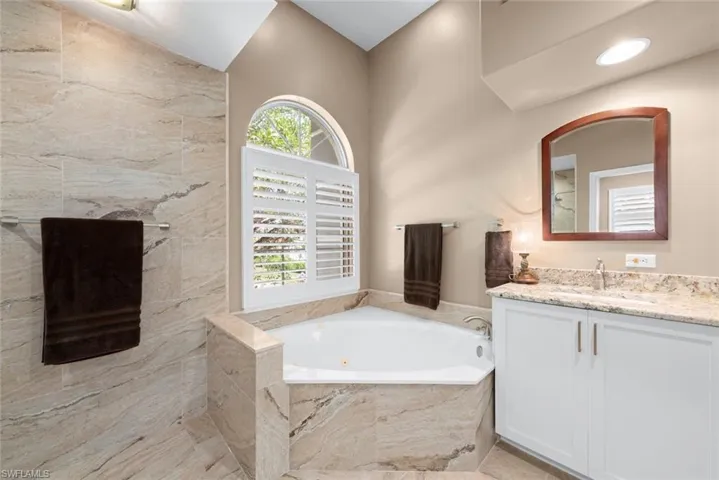 Corner soaking tub with a tiled surround, arched window featuring plantation shutters, and a vanity with a stone countertop and under-mount sink