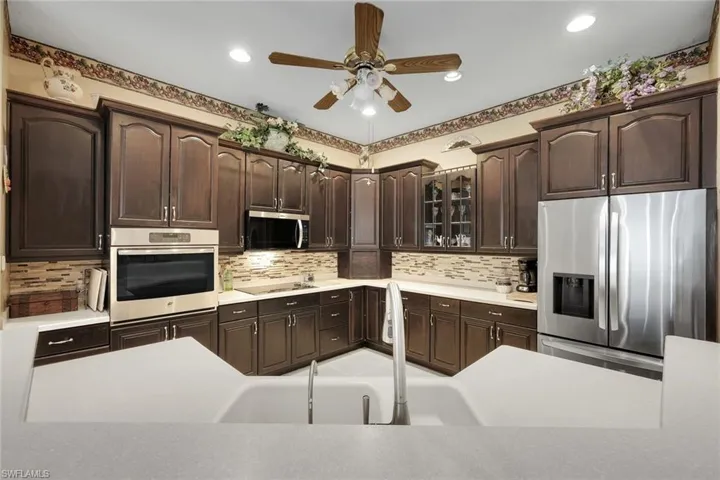 Kitchen featuring stainless steel appliances, dark wood finish cabinetry, ceiling fan, glass insert cabinets, and tasteful backsplash