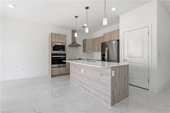 Kitchen with modern cabinets, light stone countertops, stainless steel appliances, a center island with sink, and pendant lighting