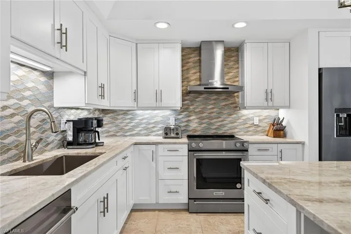Kitchen featuring stainless steel appliances, wall chimney range hood, white cabinets, light stone counters, and backsplash