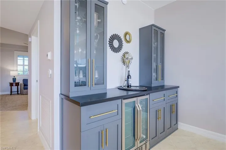 Custom dry bar featuring slate-gray cabinetry, gold-finish hardware, glass-front upper cabinets, integrated beverage refrigeration, and a dark countertop