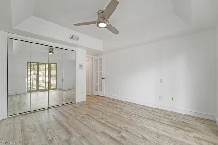 Spacious primary suite featuring gleaming wood-finish flooring, expansive mirrored closet with glass sliding doors, ceiling fan with integrated lighting, and abundant natural light from shuttered windows