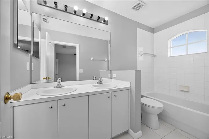 Dual vanity with integrated sinks, full-width mirror, overhead lighting fixture, and a tiled bathtub with an arched window