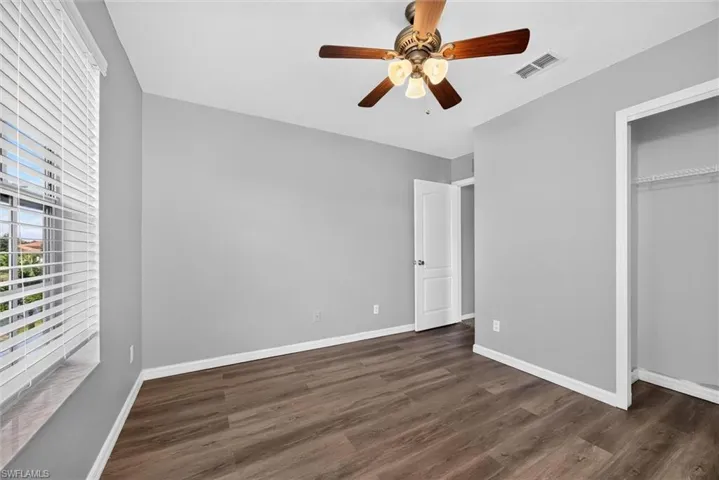 Wood-finish flooring, light gray wall paint, window with blinds, ceiling fan with integrated lighting, and a built-in closet with wire shelving