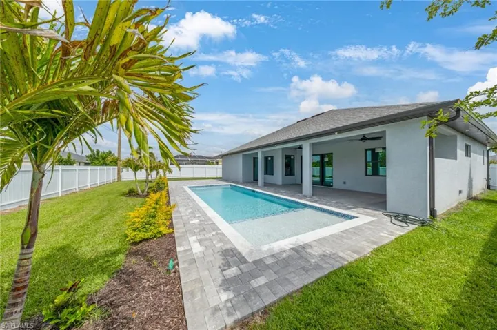 Expansive backyard featuring a rectangular swimming pool with integrated spa, paver deck, covered patio, white vinyl fencing, and mature landscaping