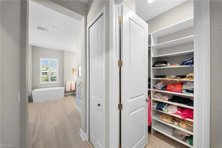 Hallway with light-colored flooring, leading to a bathroom with a freestanding white tub and a window with white shutters