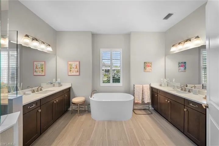 This bathroom features a freestanding bathtub, light wood-style flooring, and a window with plantation shutters