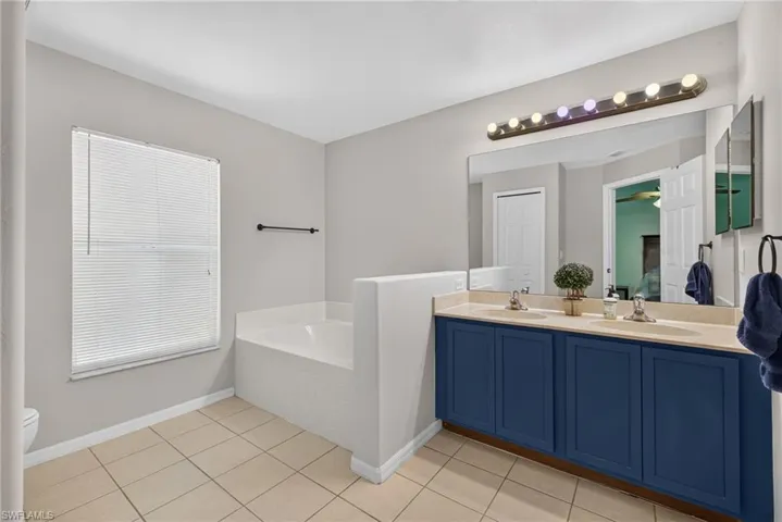 En-suite dual-sink vanity with navy blue cabinetry, solid surface countertop, large wall-mounted mirror, integrated soaking tub, and ceramic tile flooring.