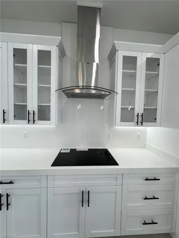 Kitchen featuring wall chimney exhaust hood, white cabinets, glass insert cabinets, black electric stovetop, and light stone countertops