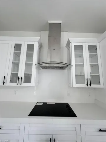Kitchen featuring glass insert cabinets, white cabinets, wall chimney range hood, black electric stovetop, and light stone counters