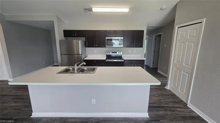 Kitchen with light countertops, appliances with stainless steel finishes, dark wood-style flooring, and a center island with sink