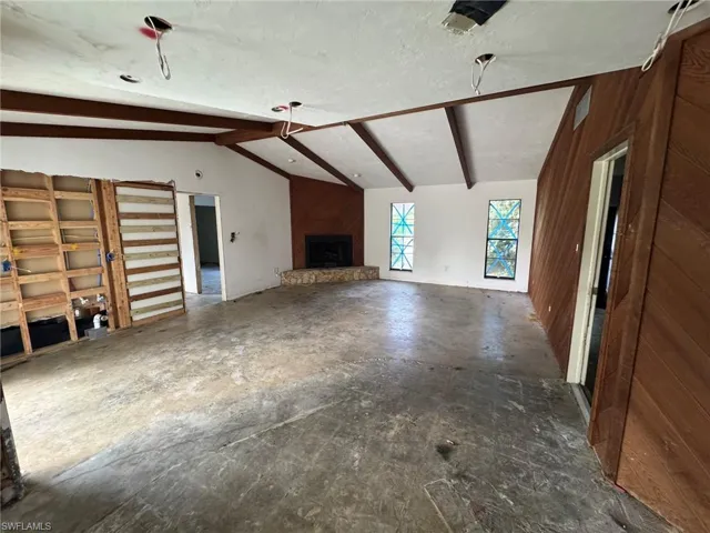 Vaulted ceiling with exposed wood beams, corner fireplace with stone surround, wood-paneled accent wall, and multiple window openings