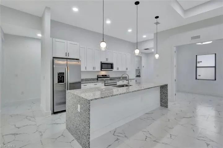 Kitchen featuring stainless steel appliances, white cabinets, light marble finish flooring, light stone countertops, and a center island with sink