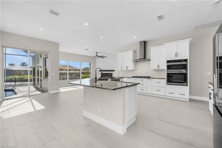 Kitchen featuring dark granite countertops, recessed lighting, wall chimney exhaust hood, a center island with sink, and white cabinets