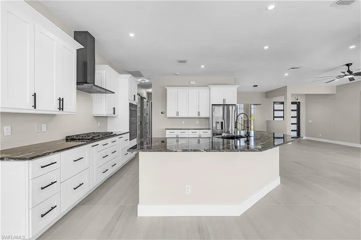 Kitchen featuring dark granite countertops, a large island, wall chimney range hood, white cabinetry, and stainless steel appliances