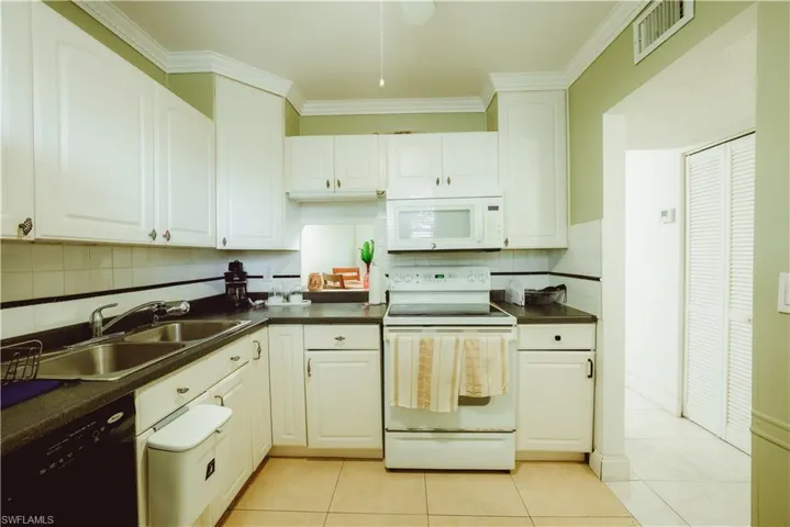 Kitchen featuring white appliances, ornamental molding, light tile patterned flooring, dark countertops, and white cabinetry