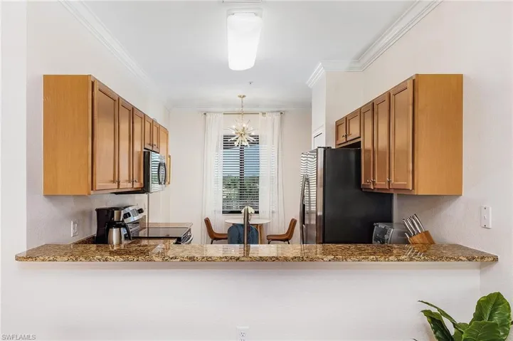 Kitchen featuring stainless steel appliances, brown cabinetry, a chandelier, crown molding, and light stone counters