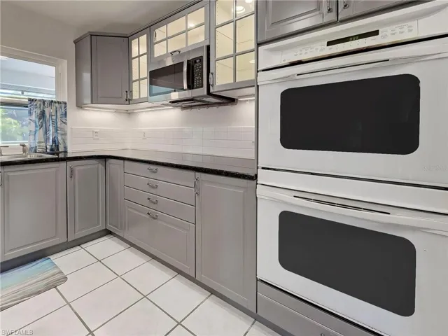 Kitchen with gray cabinetry, white double oven, dark stone countertops, stainless steel microwave, and light tile patterned floors