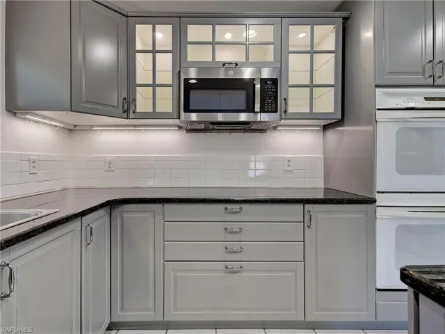 Kitchen featuring white double oven, stainless steel microwave, glass insert cabinets, dark stone counters, and gray cabinetry
