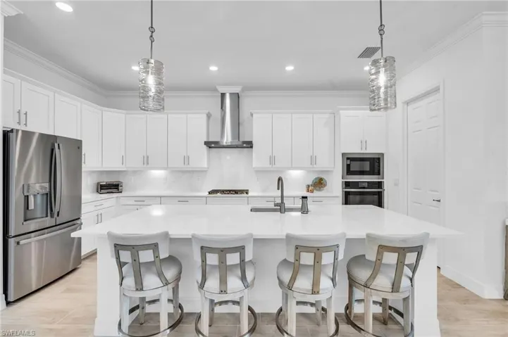 Kitchen featuring white cabinetry, stainless steel appliances, an island with sink, decorative backsplash, and wall chimney range hood