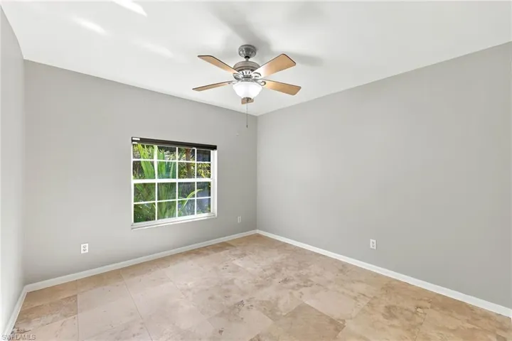 Spacious room featuring tile flooring, a window with exterior foliage, a ceiling fan with light fixture, and neutral painted walls