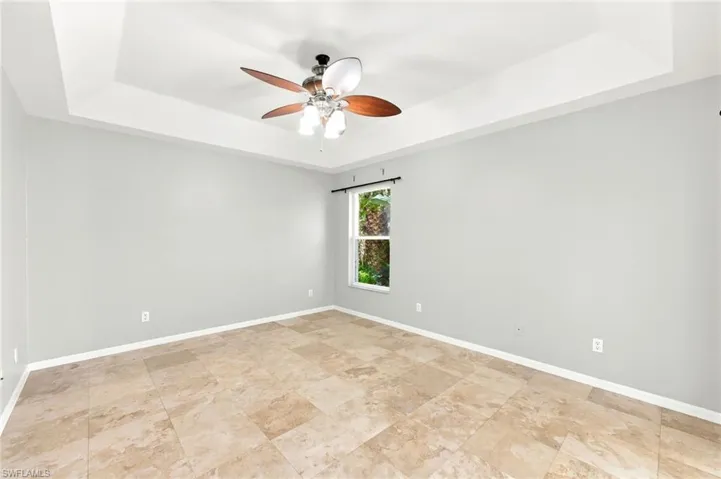 Room with tray ceiling, ceiling fan with integrated lighting, window with exterior foliage, light gray walls, and large format floor tiles