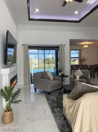 Living area featuring marble tiled flooring, a tray ceiling, a ceiling fan, a glass covered fireplace, and crown molding