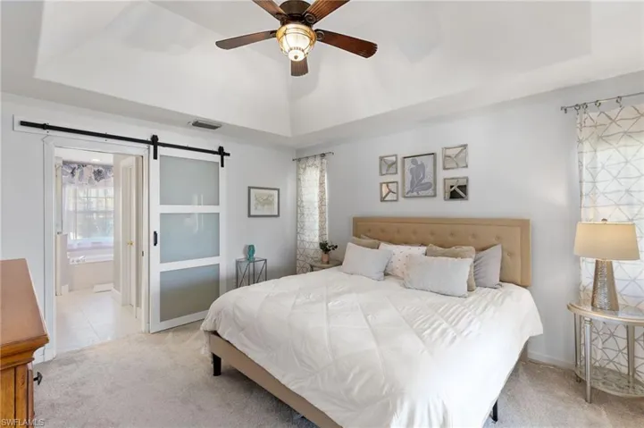 Bedroom featuring carpet, a barn door, a raised ceiling, a ceiling fan, and connected bathroom