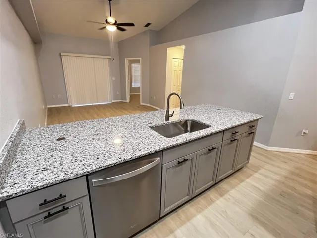 Kitchen with gray cabinetry, light wood finished floors, stainless steel dishwasher, light stone countertops, and vaulted ceiling