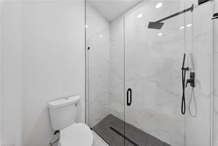 Frameless glass shower enclosure featuring large format white tile walls, a matte black rainfall showerhead, and a matching hand shower